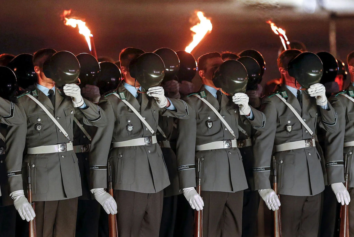 German Bundeswehr soldiers in dress uniform participate in a nighttime military ceremony, holding their helmets before their faces as ceremonial torches burn behind them.