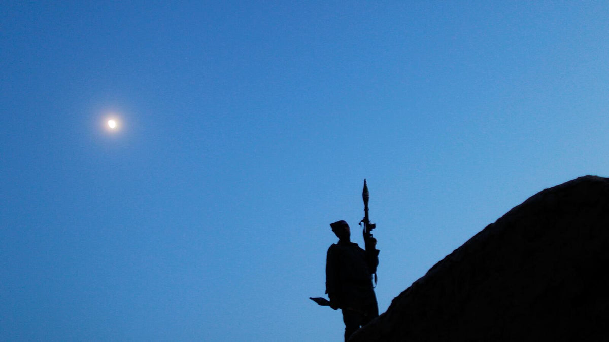 Silhouette of an armed Afghan fighter standing atop a rocky ridgeline at dusk beneath a bright moon, holding an RPG launcher against a deep blue twilight sky.