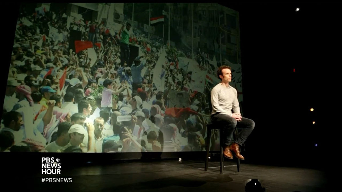 Elliot Ackerman sits on stage during a PBS NewsHour essay with a large image of Arab Spring protesters projected behind him, reflecting on war, revolution, and friendship in Syria and the broader Middle East.