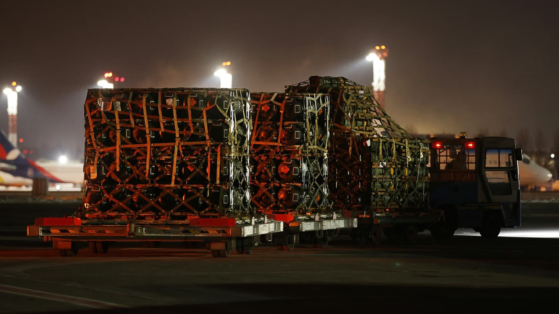Palletized military cargo secured with netting is transported across an airport tarmac at night, illuminated by floodlights as aircraft and logistics operations continue in the background.